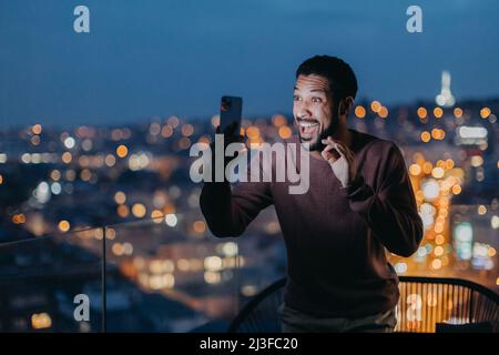 Fröhlicher junger Mann, der Selfie auf dem Balkon mit Blick auf die Stadt gemacht hat. Stockfoto