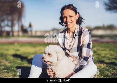 Glückliche Frau spielt mit ihrem weißen bichon-Frisehund an sonnigen Tagen im Park. Stockfoto