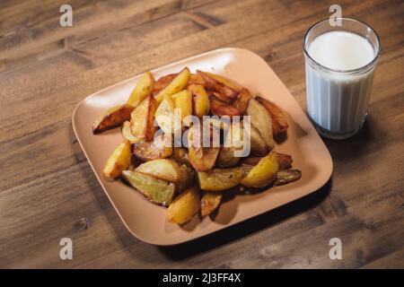 Gebackene knusprige Kartoffeln auf einem braunen Teller und Milch in einem geschnittenen Glas auf einem Holztisch. Stockfoto