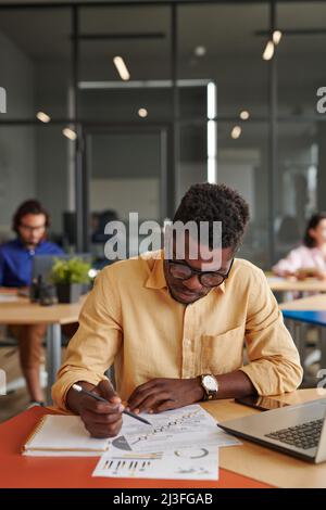 Beschäftigter junger afroamerikanischer Geschäftsmann in Brillen, der in einem gemeinsamen Arbeitsraum sitzt und das Diagramm analysiert Stockfoto