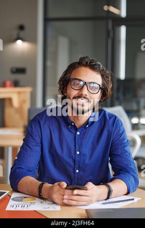 Porträt eines fröhlichen jungen Geschäftsmannes mit gemischtem Rennen und Bart in blauem Hemd mit Smartphone am Tisch im Büro Stockfoto