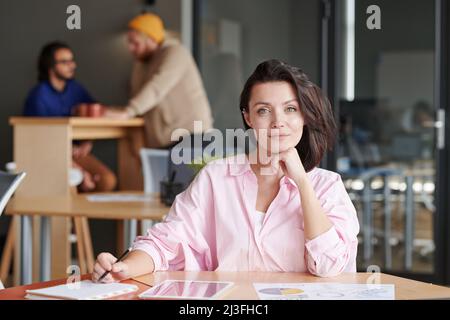 Content-Porträt Attraktive junge kaukasische Frau mit braunem Haar, die am gemeinsamen Arbeitsplatz am Tisch sitzt Stockfoto