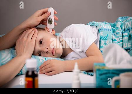 Eine Mutter misst die Temperatur in ihrem Ohr mit einem elektronischen Thermometer für ihr krankes Kind, das im Bett vor einem Tisch mit Medikamenten liegt. Fokus Stockfoto