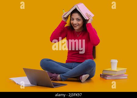 Porträt einer glücklichen jungen Frau, die die Kamera anschaut und ein Buch auf dem Kopf hält Stockfoto