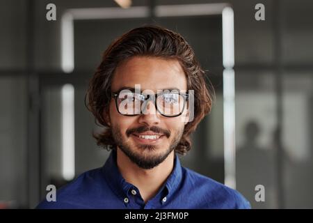 Porträt eines glücklichen jungen Mixed-Race-Managers mit Bart, der eine Brille trägt und im Büro die Kamera anlächelt Stockfoto