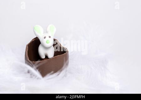 Weißer Osterhase in einem zerrissenen Schokoladenei in einem weißen, weichen Federnest, Nahaufnahme, Kopierraum. Osterkaninchen in einem zerrissenen Schokoladenei am Stockfoto