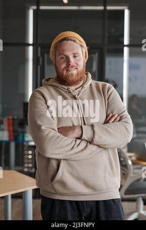Porträt eines lächelnden jungen bärtigen kaukasischen Mannes mit Hut und Kapuzenpullover, der mit gekreuzten Armen im Büro steht Stockfoto
