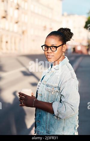 Content-Porträt eine junge schwarze Frau in einer Brille in einer Jeansjacke, die Kaffee zum Mitnehmen gegen das Stadtbild hält Stockfoto