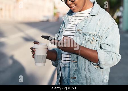 Lächelnde junge schwarze Frau in Denim-Jacke, die Kaffeetasse hält und auf der Straße telefoniert Stockfoto