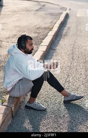 Ruhiger junger bärtiger Mann mit weißem Kapuzenpullover, der auf einem Bordstein sitzt und Smartphone benutzt, während er Musik über Kopfhörer genießt Stockfoto
