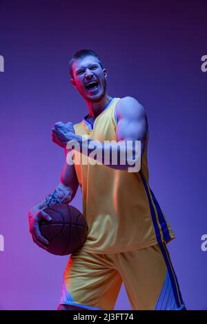 Ein aufgeregter Mann, ein Basketballspieler, der isoliert auf purpurem Hintergrund in Neonlicht schrie. Ziele, Sport, Bewegung, Aktivitätskonzepte. Stockfoto