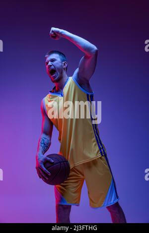 Ein aufgeregter Mann, ein Basketballspieler, der isoliert auf purpurem Hintergrund in Neonlicht schrie. Ziele, Sport, Bewegung, Aktivitätskonzepte. Stockfoto
