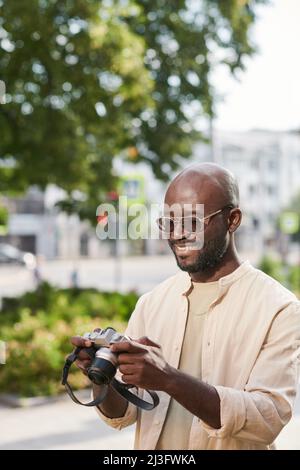 Lächelnder junger afroamerikanischer Fotograf in einer Brille, der Fotos draußen vor der Kamera durchschaut Stockfoto