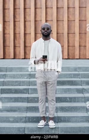 Porträt eines seriös selbstbewussten, hübschen jungen schwarzen Mannes, der mit Gadget auf der Treppe steht Stockfoto