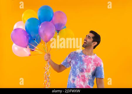 Fröhlicher junger Mann in farbenfrohem T-Shirt mit einem Haufen Ballons in der Hand Stockfoto