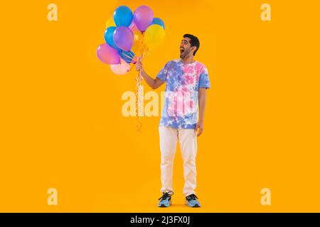 Fröhlicher junger Mann in farbenfrohem T-Shirt mit einem Haufen Ballons in der Hand Stockfoto