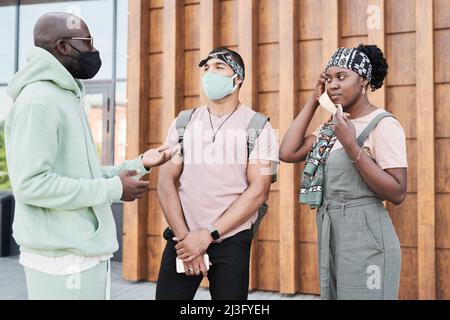 Junge afroamerikanische Frau im Kopftuch trägt eine Maske, während sie sich mit Freunden im modernen Gebäude trifft Stockfoto