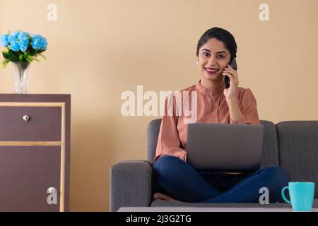 Glückliche indische Frau im Gespräch auf dem Smartphone während der Arbeit von mit Laptop Stockfoto