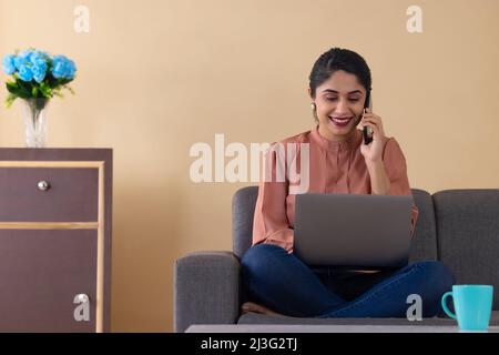 Glückliche indische Frau im Gespräch auf dem Smartphone während der Arbeit von mit Laptop Stockfoto