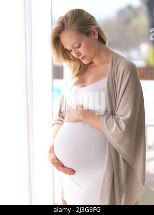 So viel Zuneigung für ihr kleines Brötchen im Ofen. Kurzer Schuss einer attraktiven jungen Schwangeren, die ihren Bauch hält, während sie in ihrem Haus steht. Stockfoto