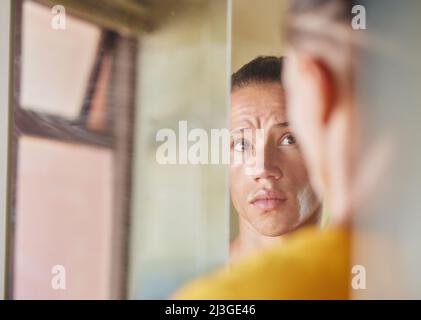 Glühend und bereit, für den Tag zu gehen. Aufnahme eines jungen Mannes, der zu Hause in seinem Badezimmer in der Spiegelung seines Spiegels auf sein Gesicht schaut. Stockfoto