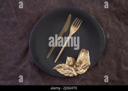 Goldenes Besteck und Blätter auf einer schwarzen Keramikplatte, auf braunem Lederhintergrund Stockfoto