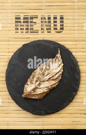 Menüwort auf einem Bambus-Tischset, mit einer Steinplatte und einem goldenen Blatt Stockfoto