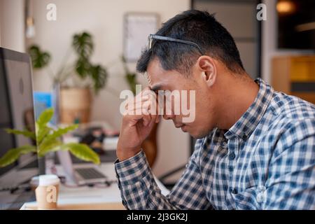 Ein junger asiatischer Geschäftsmann reibt sich die Augen, während er versucht, an seinem Schreibtisch zu arbeiten Stockfoto