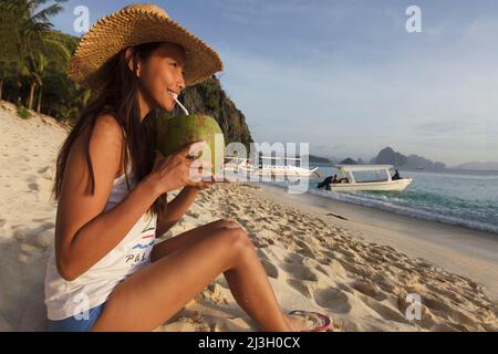 Philippinen, Palawan, El Nido, Bacuit-Archipel, Seven Commandos Beach, Eine junge philippinische Frau sitzt am Strand und trinkt aus einer Kokosnuss mit einem Stroh Stockfoto
