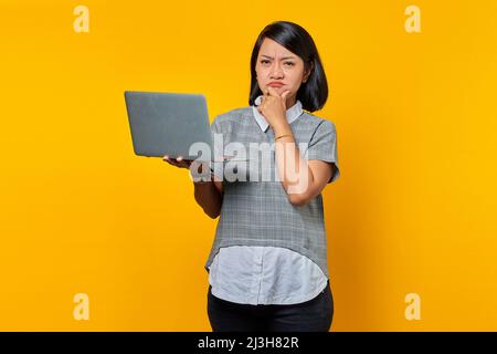 Porträt einer schönen asiatischen Frau, die ihren Laptop hält und mit der Hand am Kinn über etwas nachdenkt und die Kamera auf gelbem Hintergrund anschaut Stockfoto