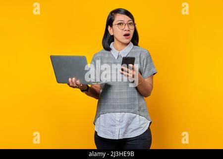 Schockiert junge schöne Frau hält Smartphone und Laptop Blick beiseite auf gelben Hintergrund Stockfoto