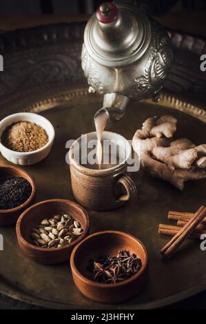 Chai-Tee-Zutaten auf einem Messingteller mit Teekocher, Zimt, schwarzem Pfeffer, Ingwer, Muskatnuss, Kardamom, Sternanis, Nelken, schwarzer Tee, Milch und brauner Zucker Stockfoto