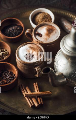 Chai-Tee-Zutaten auf einem Messingteller mit Teekocher, Zimt, schwarzem Pfeffer, Ingwer, Muskatnuss, Kardamom, Sternanis, Nelken, schwarzer Tee, Milch und brauner Zucker Stockfoto