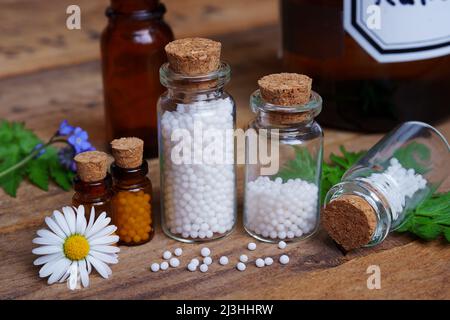 Medizinische Glasfläschchen auf Holztisch, gefüllt mit Globulis. Dekorieren Sie mit Blättern und Blumen. Stockfoto