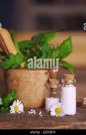 Medizinische Glasfläschchen auf Holztisch, gefüllt mit Globulis. Dekorieren Sie mit Blättern und Blumen. Stockfoto