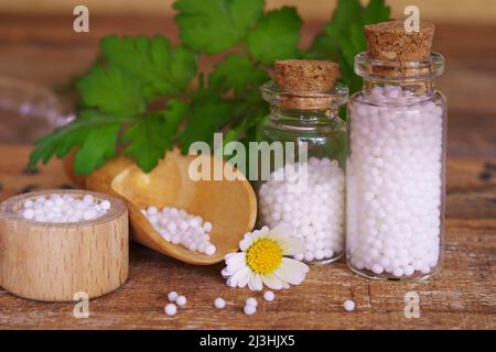 Medizinische Glasfläschchen und Schalen auf Holztisch, gefüllt mit Globulis. Mit frischen grünen Blättern dekorieren. Stockfoto