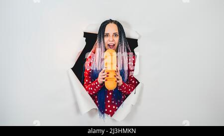 Junge Frau beißt weißes Brot, steht in einem Loch mit weißem Hintergrund. Hungrige Frau, die im Studio Brot isst Stockfoto