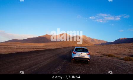 Spanien, Kanarische Inseln, Fuerteventura, Südwestspitze, karge Landschaft, Silbernes Auto auf unbefestigten Straßen, rotbraune Lavahügel im Hintergrund, himmelblau mit wenigen Wolken Stockfoto