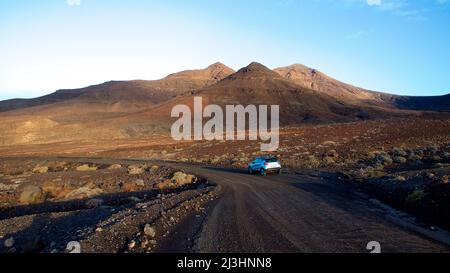 Spanien, Kanarische Inseln, Fuerteventura, Südwestspitze, karge Landschaft, Unbefestigte Straße, einzelnes silbernes Auto auf unbefestigte Straße, Hügel im Hintergrund, himmelblau Stockfoto