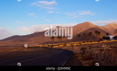 Spanien, Kanarische Inseln, Fuerteventura, Südwestspitze, karge Landschaft, Unbefestigte Straße, Palmen, Einzelgebäude, Wohnmobil neben unbefestigten Straße, hellblau mit grau-weißen Wolken Stockfoto