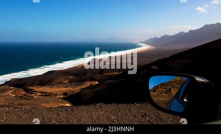 Spanien, Kanarische Inseln, Fuerteventura, südwestliche Spitze, karge Landschaft, Blick von oben auf den einsamen Nordteil der Südspitze, rechter Wagenspiegel im Bild, Gebirgskette im Hintergrund, Meer mit starker Brandung, einsamer Strand Stockfoto