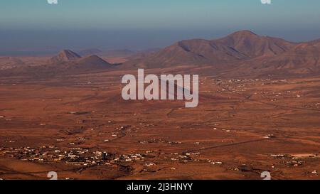 Spanien, Kanarische Inseln, Fuerteventura, Mirador Morro Velosa, Blick nach Norden auf eine karge braune Landschaft mit isolierten vulkanischen Hügeln, kleinen Dörfern und isolierten Gebäuden, Meer im Hintergrund, himmelblau Stockfoto