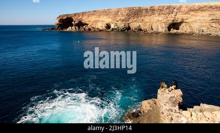 Spanien, Kanarische Inseln, Fuerteventura, Westküste, Ajuy, bucht von Ajuy, felsige Küste im Hintergrund, Einzelperson, die auf einem felsigen Ausbisse sitzt und das rauschende Meer anschaut Stockfoto