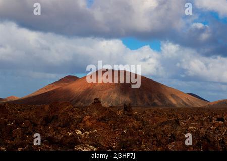 Kanarische Inseln, Lanzarote, Vulkaninsel, Nationalpark Timanfaya, Vulkanlandschaften, Blick von der Straße in den Nationalpark, roter Lavagestein im Vordergrund, rotbraune Vulkankrater in der Mitte, blauer Himmel mit grau-weißen Wolken, der Krater ist im Bild groß Stockfoto