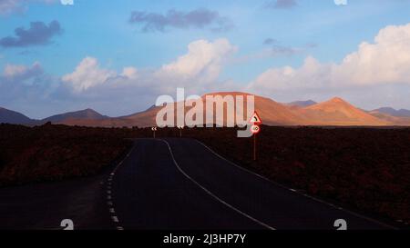 Kanarische Inseln, Lanzarote, Vulkaninsel, Nationalpark Timanfaya, Vulkanlandschaften, Eine asphaltierte Straße im Schatten führt zum Nationalpark, rot-brauner Vulkankrater in der Mitte, blauer Himmel, grau-weiße Wolken, Verkehrszeichen '40', Verkehrszeichen 'scharfe linke Kurve' Stockfoto