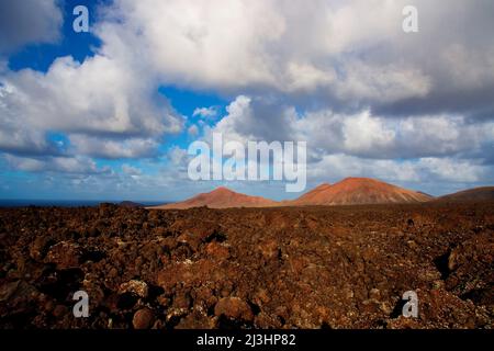 Kanarische Inseln, Lanzarote, Vulkaninsel, Nationalpark Timanfaya, Vulkanlandschaften, Blick von der Straße in den Nationalpark, roter Lavagestein im Vordergrund, rotbraune Vulkankrater in der Mitte, blauer Himmel mit grau-weißen Wolken, zwei rote Vulkankrater nebeneinander in der Mitte Stockfoto