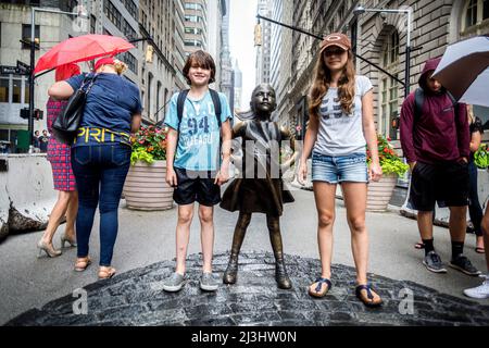 Finanzdistrikt, New York City, NY, USA, 14 Jahre altes kaukasisches Teenager-Mädchen und 12 Jahre altes kaukasisches Teenager-Mädchen – beide mit braunen Haaren und sommerlichem Styling neben der Statue „Fearless Girl“ von Kristen Visbal in der Broad Street vor der New Yorker Börse Stockfoto
