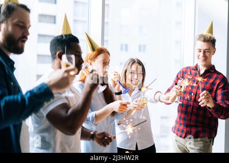 Erfolgreiches Business-Team aus jungen, multiethnischen Geschäftsleuten, die bei einer Firmenfeier bengalische Lichter und knallende Gläser Champagner halten. Stockfoto