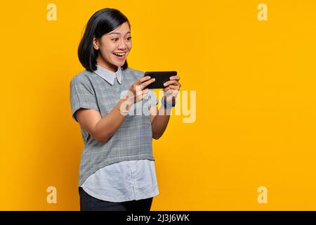 Porträt einer fröhlichen jungen asiatischen Frau, die Videospiel auf dem Mobiltelefon auf gelbem Hintergrund spielt Stockfoto