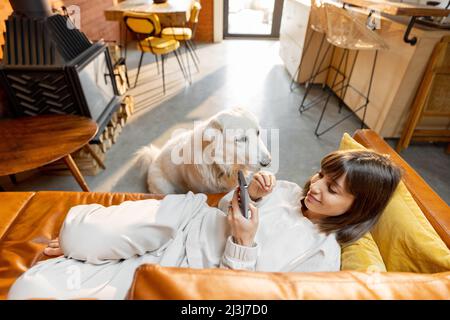 Die Frau lag entspannt auf der Couch und telefoniert und verbrachte zu Hause Zeit mit dem Hund Stockfoto